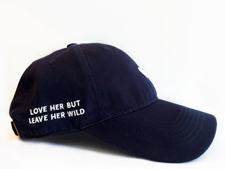 Navy blue cotton baseball cap with a white heart emblem embroidered by Atticus Poetry and "Love Her But Leave Her wild" saying embroidered
